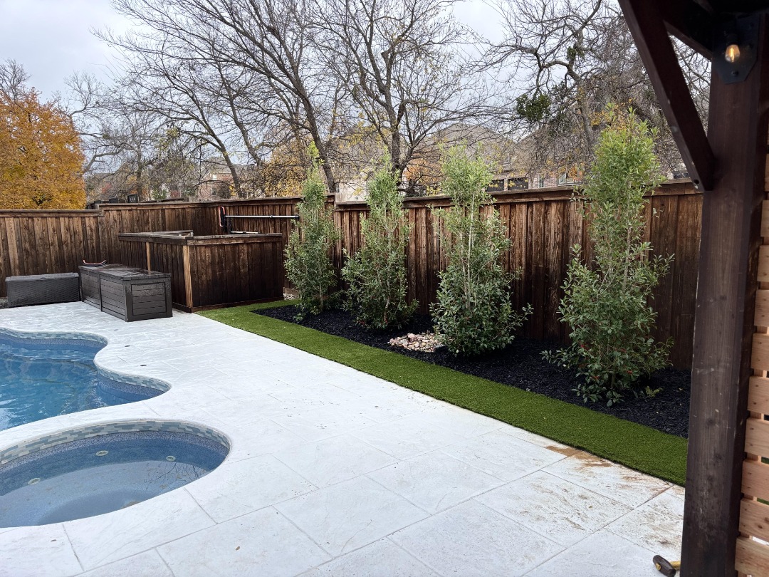 Backyard poolside features white concrete deck, green privacy shrubs, dark mulch, and a wood fence under overcast skies.