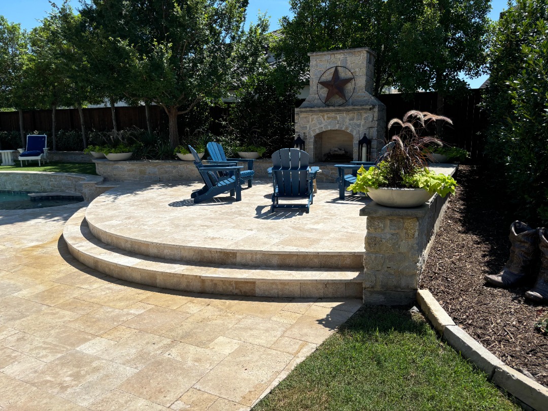 Elevated stone patio with blue Adirondack chairs surrounds a rustic fireplace, bordered by planters and lush garden landscaping.