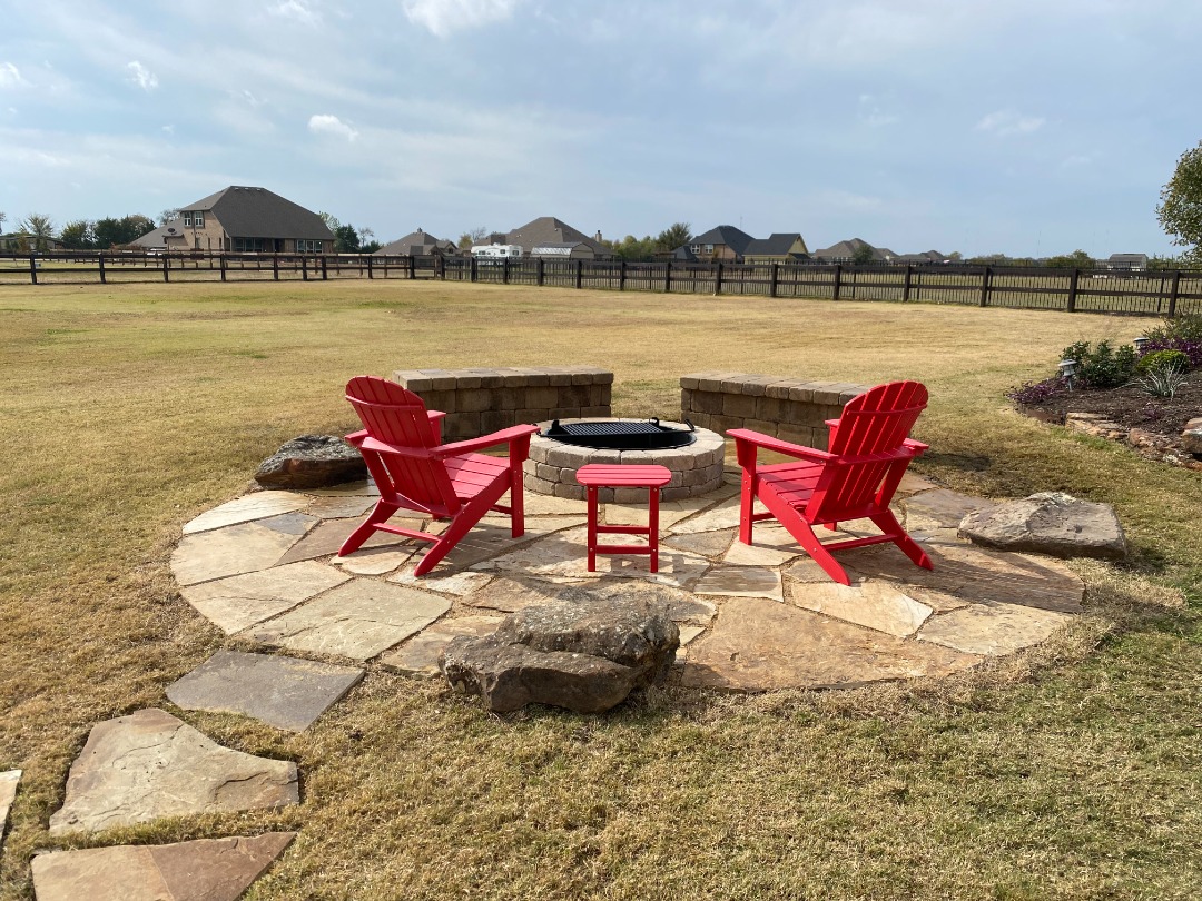 Stone patio with red Adirondack chairs surrounds a circular fire pit in a large open backyard with distant homes and fencing.