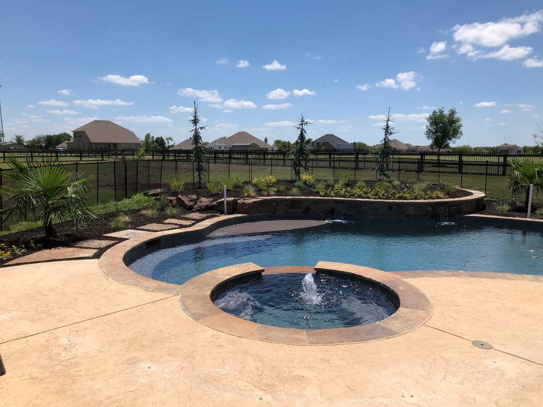 Curved pool with a small splash pool that is surrounded by tiered landscaping, palm trees, and black metal fencing.