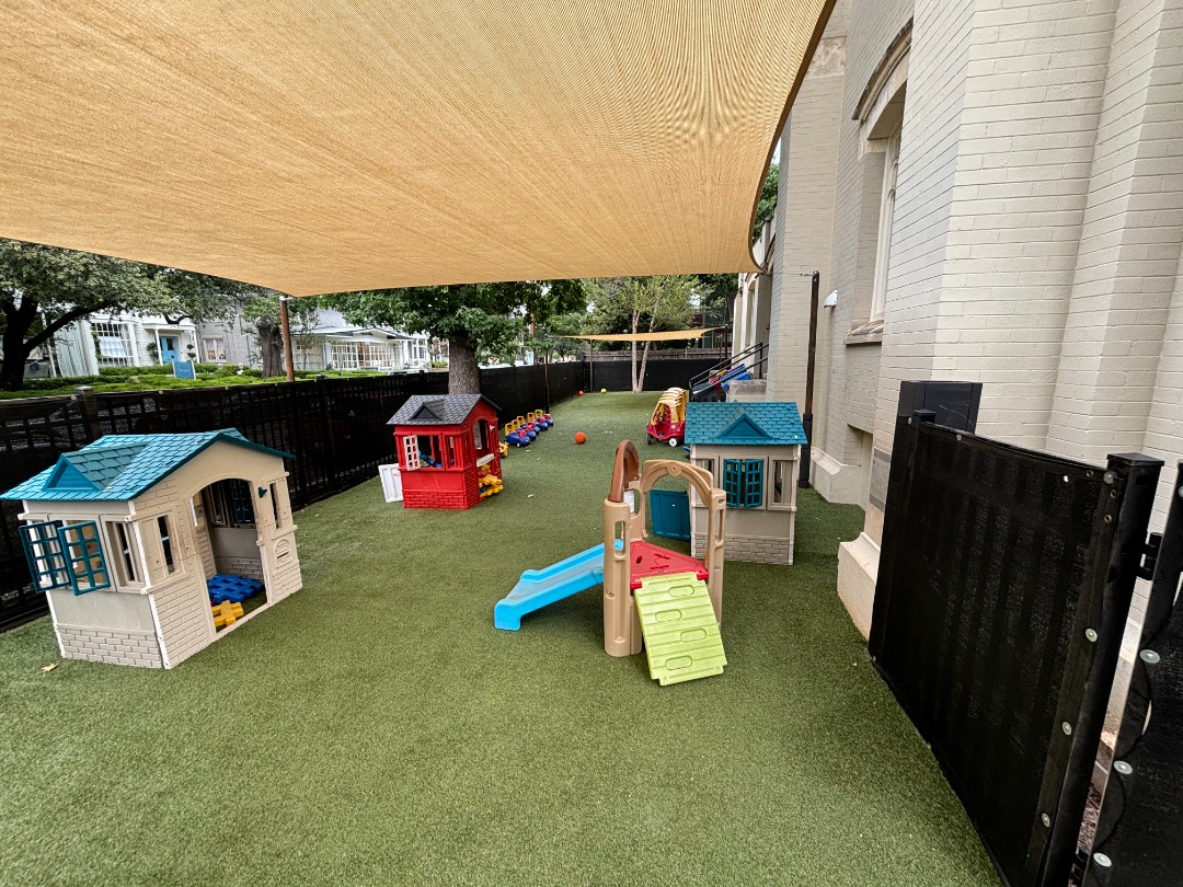 Artificial turf playground features plastic playhouses and a slide beneath a large sunshade beside a brick school building.